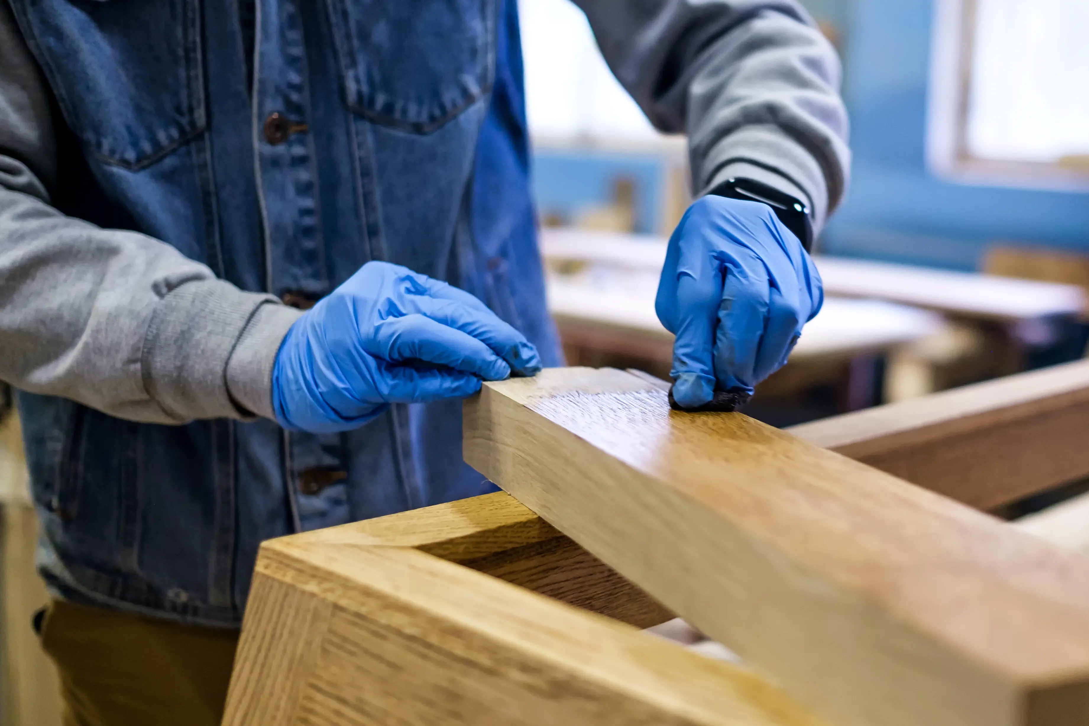 Eine Hand in blauen Schutzhandschuhen streicht eine Holzoberfläche mit brauner Farbe.